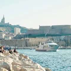 Widok na port w Marsylii z łodzią, fortecą i bazyliką na wzgórzu.