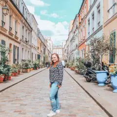Urocza ulica w 16. dzielnicy Paryża, z kolorowymi kamienicami, mnóstwem zieleni w donicach i uśmiechniętą kobietą.