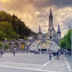 Bazylika w Lourdes z placem i pielgrzymami pod zachmurzonym niebem.