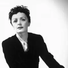 Edith Piaf w czarno-białym portrecie, w czarnym żakiecie i z charakterystyczną fryzurą.