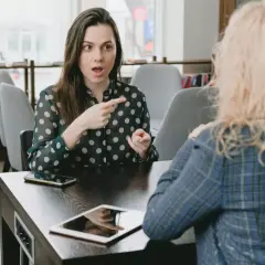 Dwie kobiety rozmawiają przy stole, jedna wskazuje palcem.