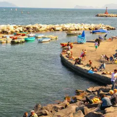 Słoneczna neapol plaża z łodziami i ludźmi odpoczywającymi na kamieniach i molo.