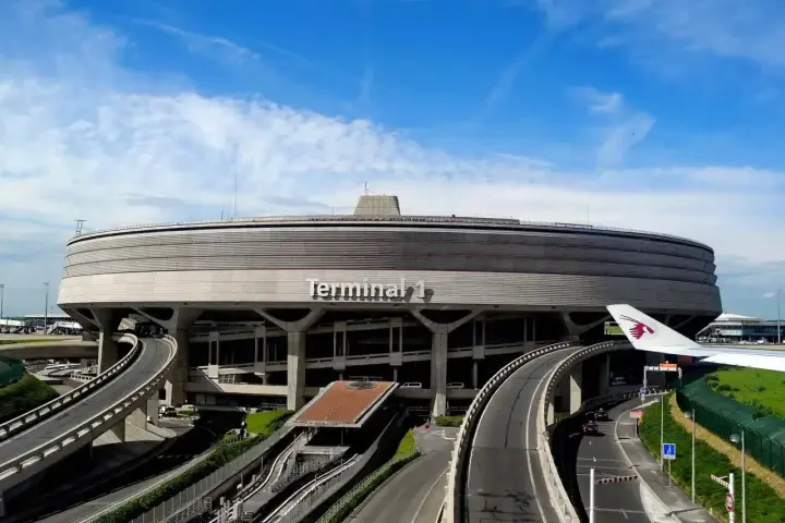 Terminal 1 na lotniskach w Paryżu, z charakterystyczną, okrągłą architekturą i drogami prowadzącymi do niego. Widoczny fragment skrzydła samolotu Qatar Airways.