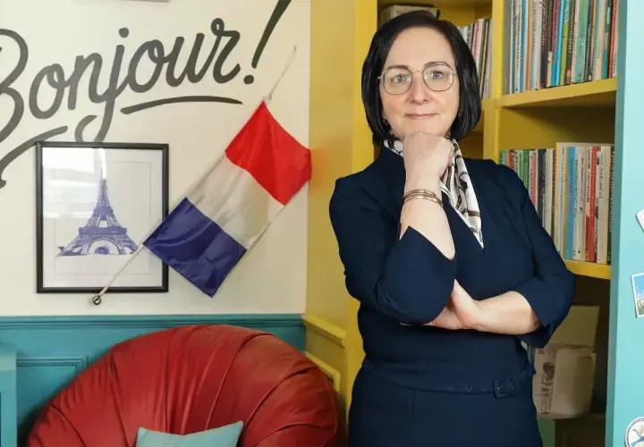 Kobieta w okularach stoi przed ścianą z napisem "Bonjour!", francuską flagą i wieżą Eiffla.
