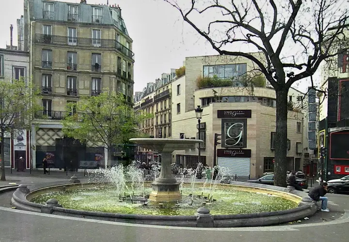 Fontanna na placu Pigalle w Paryżu, otoczona kamienicami i drzewem.