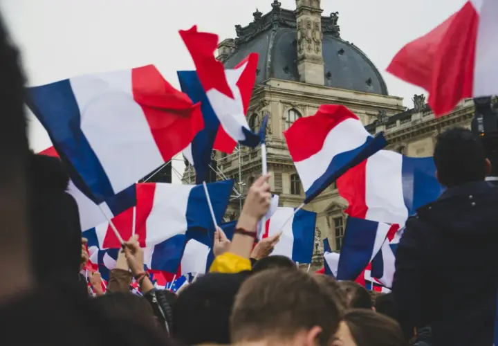 Tłum ludzi trzyma flagi francuskie. Widać charakterystyczne biało-niebiesko-czerwone pasy, jak wygląda flaga francuska.