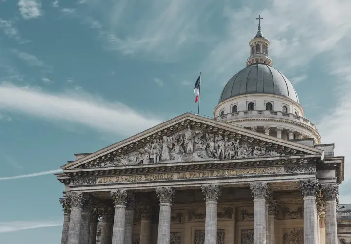 Majestatyczny Panteon w Paryżu z kopułą i francuską flagą na tle błękitnego nieba.