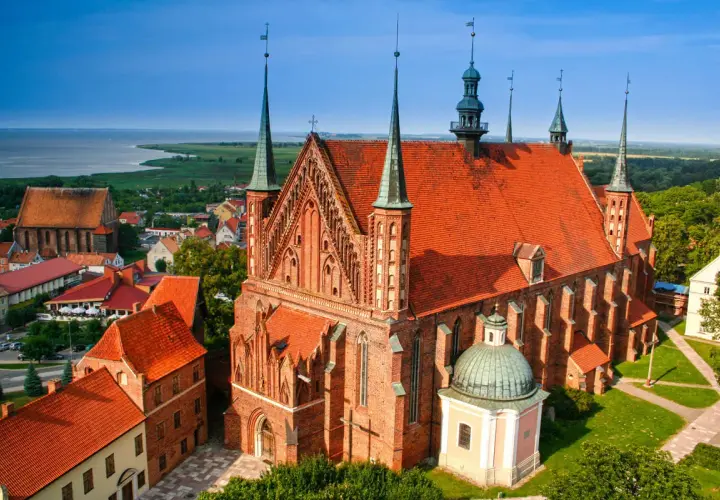 Widok na Frombork z zamkiem i katedrą. Czerwone dachy budowli kontrastują z błękitem nieba i zielenią otoczenia.