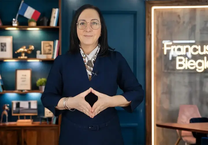 Kobieta w okularach i granatowej sukience, z rękami złożonymi w romb, na tle regału z książkami i neonu z napisem "Francuski Biegle".