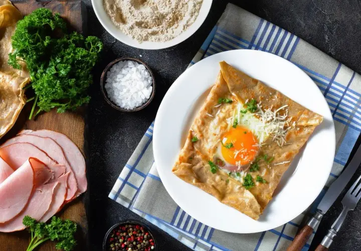 Francuska crepe z jajkiem sadzonym, serem i natką pietruszki na talerzu, obok szynka i składniki.