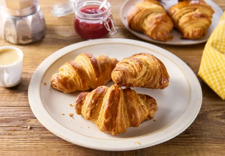 Talerz z trzema złotymi croissantami, filiżanka kawy, słoiczek dżemu i kawiarka.