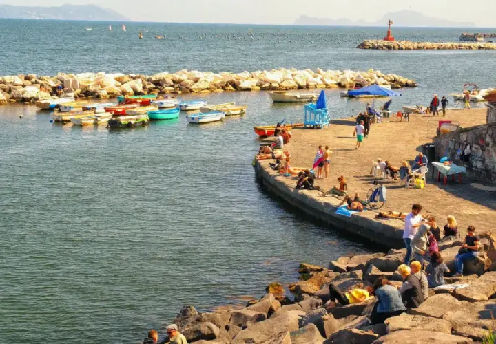Słoneczna neapol plaża z łodziami i ludźmi odpoczywającymi na kamieniach i molo.