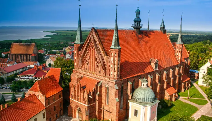 Widok na Frombork z zamkiem i katedrą. Czerwone dachy budowli kontrastują z błękitem nieba i zielenią otoczenia.