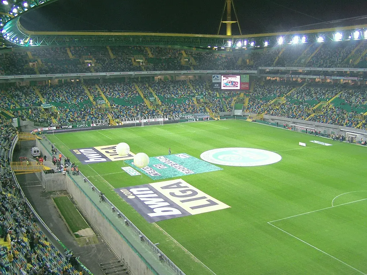 Widok na pełen widzów **sporting lizbona stadion** przed meczem. Na murawie widoczne są logotypy sponsorów i wielkie balony.