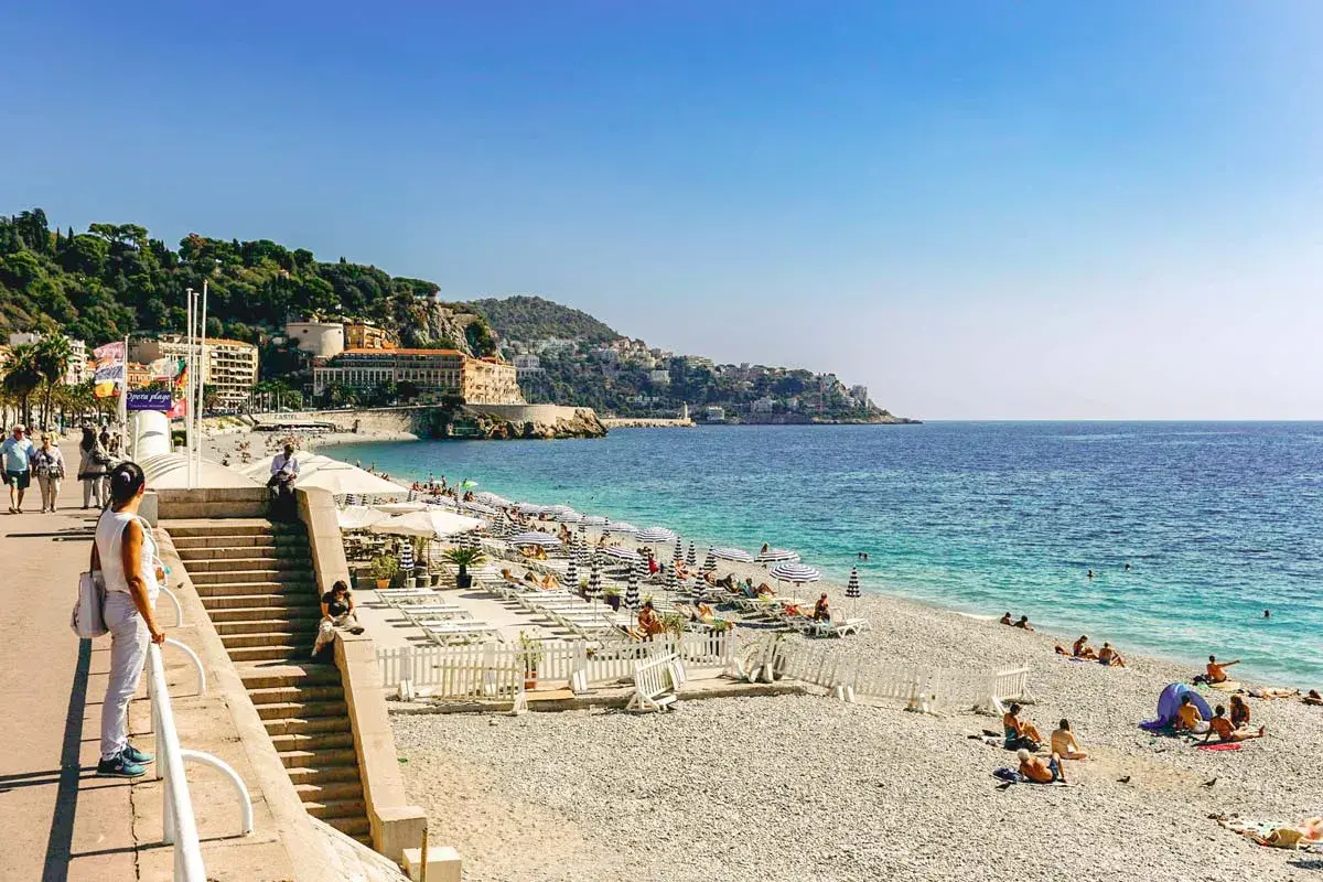 Kamienista plaża w Nicei z leżakami, parasolami i turystami.