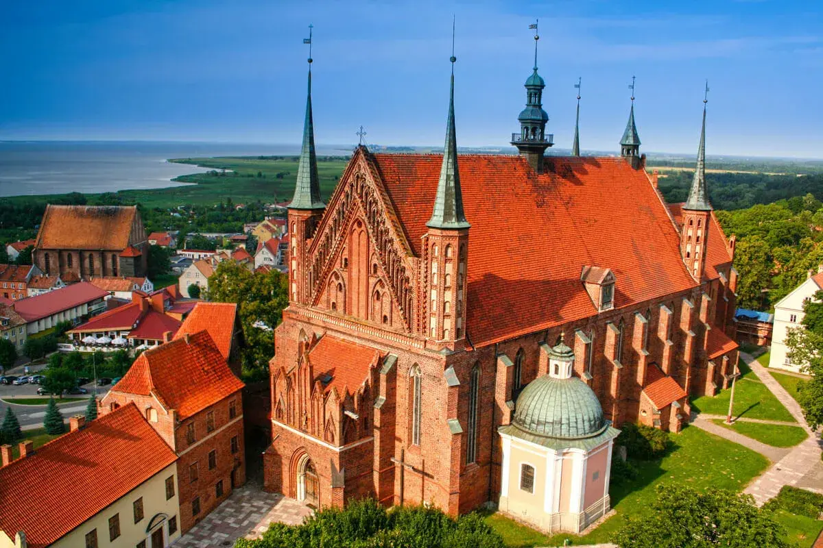 Widok na Frombork z zamkiem i katedrą. Czerwone dachy budowli kontrastują z błękitem nieba i zielenią otoczenia.