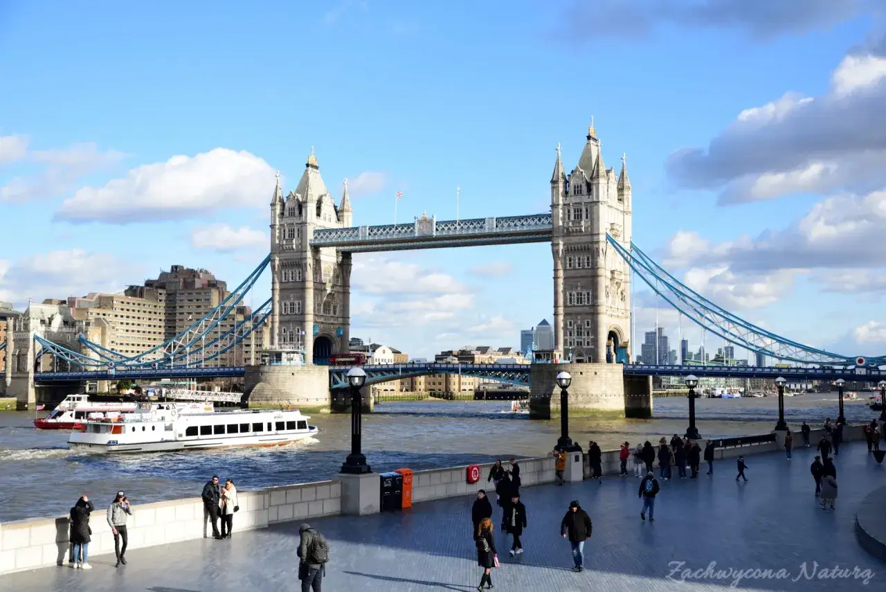 Tower Bridge w Londynie, z łodziami na Tamizie i spacerującymi ludźmi. Ciekawe wieże i niebieskie liny mostu.
