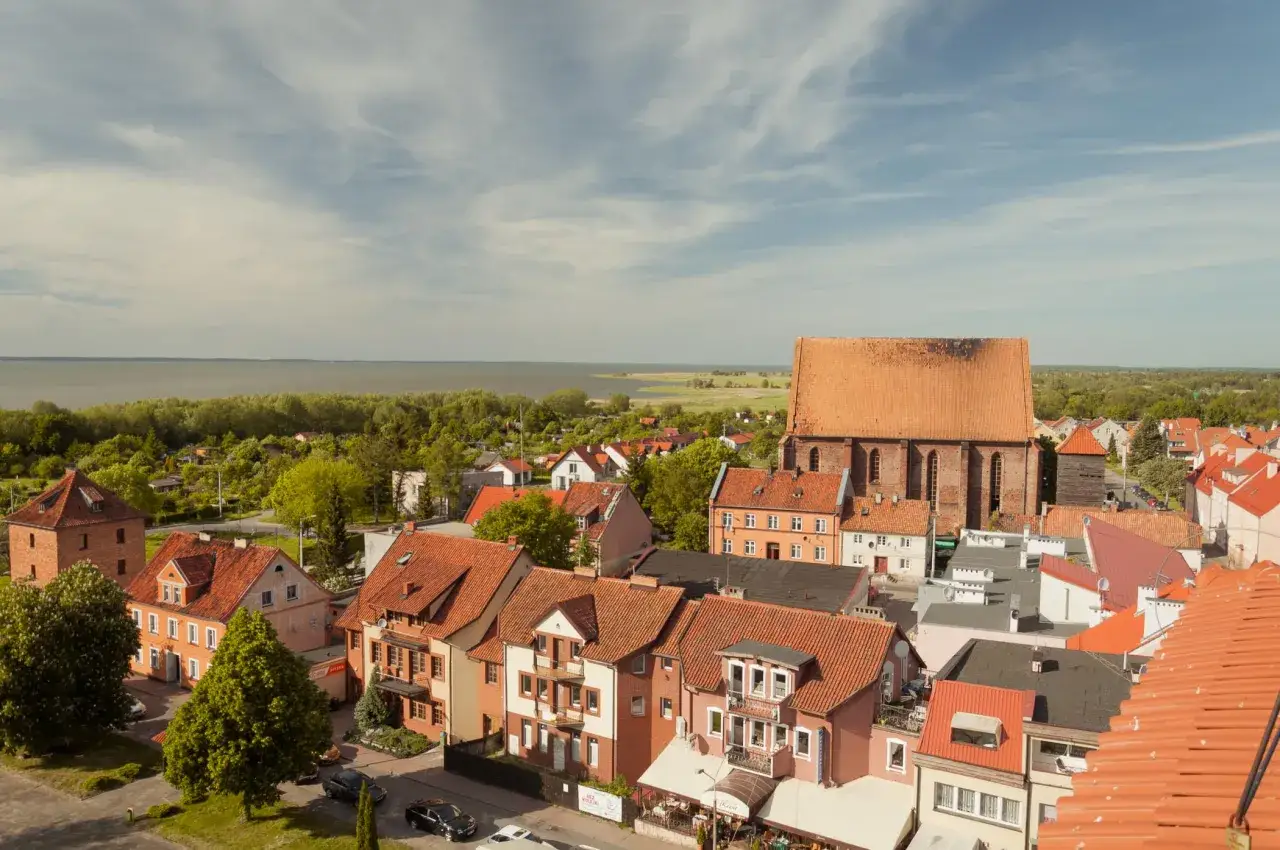 Widok na Frombork z zamkiem w tle, nad zatoką. Czerwone dachy kamienic i kościoła tworzą malowniczy krajobraz.